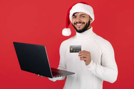 Happy hipster man wears santa claus hat holding notebook and gift box over red background. Christmas concept.の写真素材