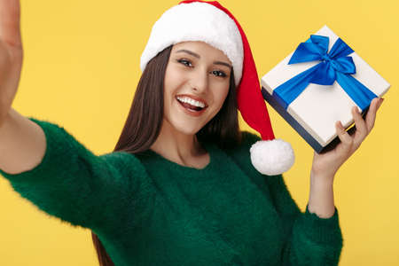 Gladful young brunette woman wear santa claus hat and green pullover take selfie with gift box over...