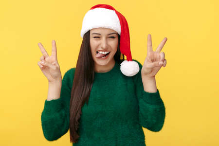 Funny young brunette woman wear santa claus hat show heart gesture to camera over yellow background.の写真素材