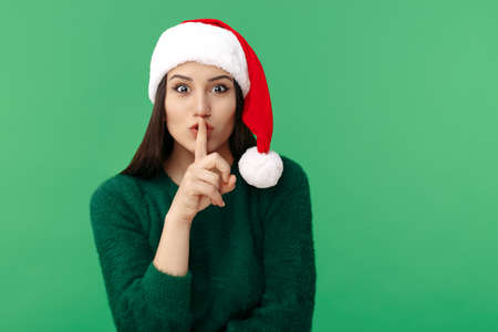 Pretty young brunette woman wear santa claus hat showing keep silent gesture over green background.の写真素材