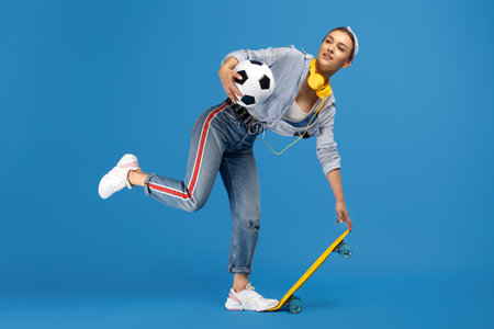Cheerful young woman with yellow penny or skateboard, soccer ball and headphones posing over blue...
