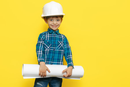 Cheerful little kid boy engineer or architect in a protective helmet holding construction plan over yellow background.の写真素材