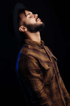 Handsome young bearded hipster in brown shirt posing with closed eyes over black background.の写真素材