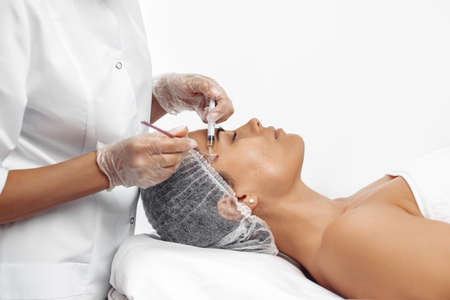 Cosmetologist applying cream on female face with brush in cosmetology salon.
