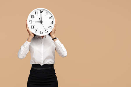 Photo of young modern business woman hold a clock in front of the face over beige background.