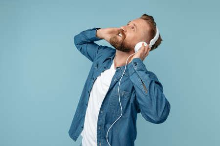 Stylish bearded blonde hipster man in jeans shirt wearing headphones over blue background.の写真素材