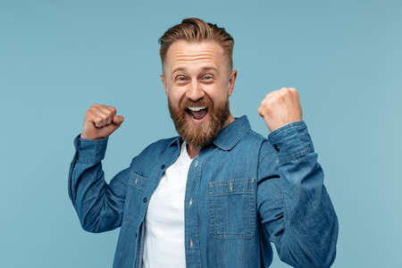 Happy bearded hipster blonde man in jeans shirt celebrate success over blue background. Look in...