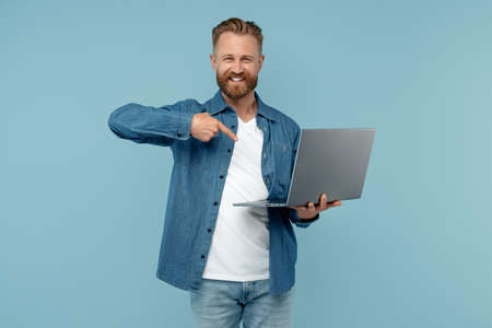 Smiling bearded blonde hipster young man in jeans shirt pointing finger on computer over blue background.の写真素材