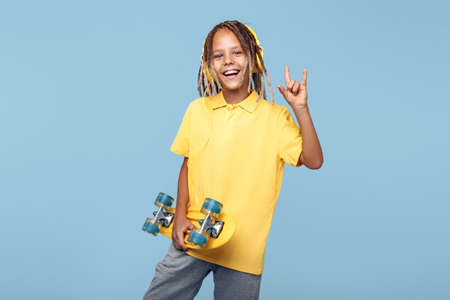 Happy little boy with african dreads with skateboard on the blue background showing rock gesture.