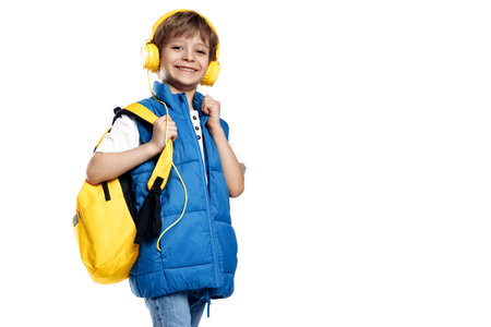Cheerful little boy wearing headphone and yellow backpack looking to camera on white background. Back to School.の写真素材