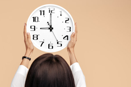 Close-up photo of young modern business woman hold a clock over head on beige background.