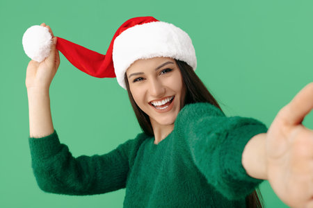 Smiling positive young brunette woman wear santa claus hat and green pullover take selfie over green background.の写真素材