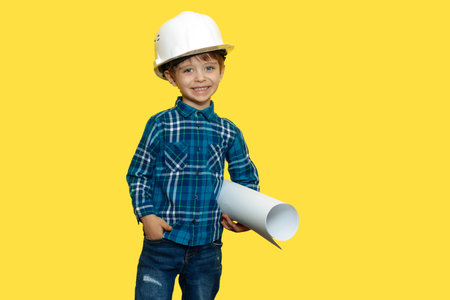 Joyful little kid boy engineer or architect in a protective helmet holding construction plan over...