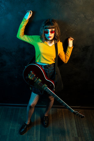 Stylish young hipster woman with curly hair with red guitar in neon lights. Rock musician posing...