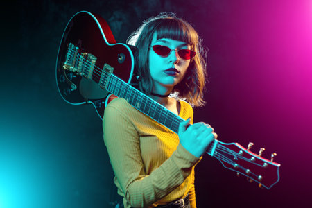 Trendy young hipster woman with curly hair with red guitar in neon lights. Rock musician posing with electrical guitar.の写真素材