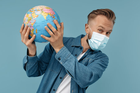 Bearded blonde hipster man in medical mask holding world globe over the head on blue background