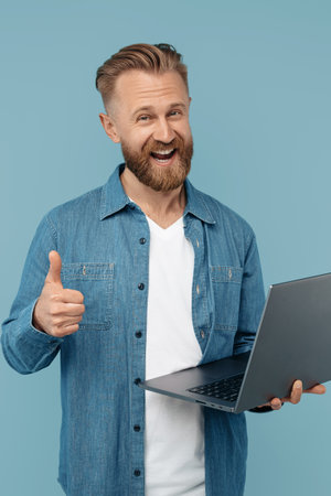 Smiling bearded blonde hipster young man in jeans shirt with laptop showing thumb up over head on blue backgroundの写真素材