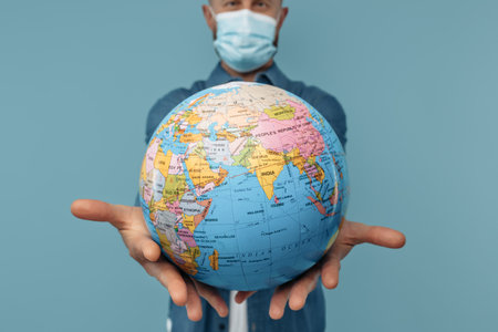 Close-up image of bearded blonde hipster man in medical mask holding world globe on blue background.の写真素材