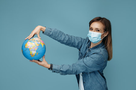Charming young woman in medical mask holding world globe over blue backgroundの写真素材