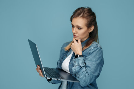 Charming young woman in jeans shirt using computer or laptop over blue backgroundの写真素材