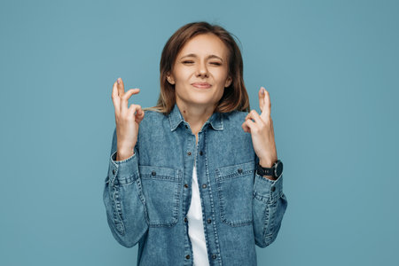 Hopeful woman holding hands with fingers crossed in hope over blue background. Woman with closed...