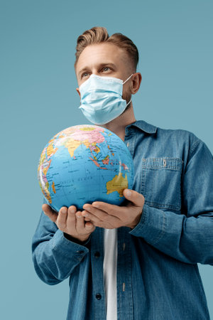 Bearded blonde hipster man in medical mask holding world globe on blue background.
