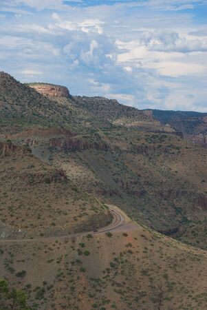 Huge desert cliffs loom over a winding highway road with swirling white clouds and a hint of blue skyの写真素材