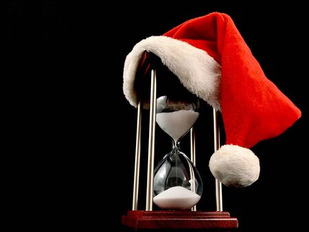Hourglass in the cap of Santa Claus. Christmas hat on glass hourglass. Glass device for measuring time, on a black background. Concept: outgoing year, new yearの写真素材