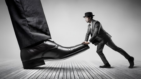 The monochrome tone emphasizes the contrast and symbolic struggle. A surreal image of a man in a suit and hat casually bending over and holding a massive foot to prevent it from stepping on him.の素材
