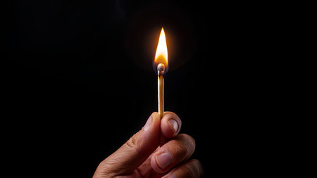 This image symbolizes potential, ignition, and the spark of ideas or beginnings. A hand holds a lit match against a completely black background, illuminating the fingers and creating a dramatic contrast.の素材