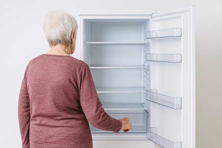 Elderly woman checks empty refrigerator in a modern kitchen while preparing for grocery shopping during the dayの素材
