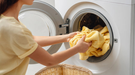 Woman loading yellow towels into a washing machine in a laundry room on a bright afternoon, preparing for a cleaning session with fresh laundryの素材