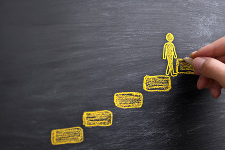 Creating a chalk drawing of a person climbing stairs on a blackboard, symbolizing progress and learning, with a hand holding chalk in a modern educational spaceの素材
