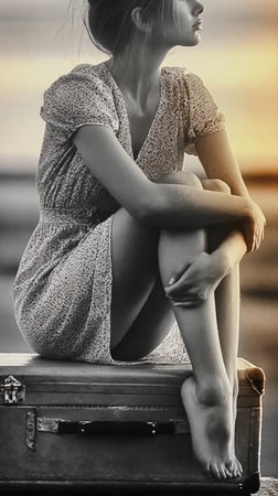 Woman sitting on a suitcase by the shore, reflecting at sunset in a tranquil and serene coastal sceneの素材
