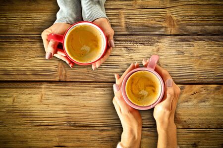 Young female manicured hands holding cups of coffee with fresh foam on dark rustic wooden table background. Free space for your decoration, copy space, top view, flat photo. Cold winter Christmas time.の写真素材