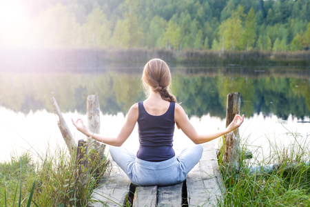 A beautiful woman sits in a pose of half lotus on a wooden pier on the shore of the lake, a stunning view of nature, she practices yoga meditation. The energy of Kundaliniの写真素材