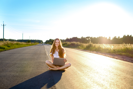 Beautiful light-haired girl sitting with laptop and smiling. On the road going beyond the horizon at sunset.の写真素材