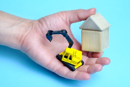 Toy yellow crawler excavator and house from wooden childrens cubes in male hand isolated on white background. Construction equipment. Buildingの写真素材