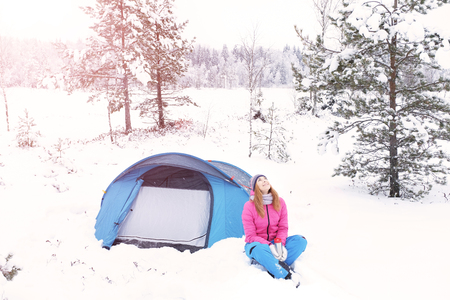 Happy smiling red-haired young girl with a  near the blue tourist tent on the background of the winter forest.の写真素材