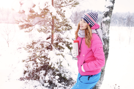 Delightful happy young red-haired girl in a winter sports suit, having fun in the winter forest. Drinking hot tea from a  cup against the backdrop of a winter forest.の写真素材