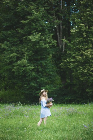 Beautiful girl with basket of wild flowers. Woman with blond curly hair with a bouquet in a straw hat outdoors.の写真素材