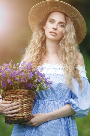 Beautiful girl with basket of wild flowers. Woman with blond curly hair with a bouquet in a straw hat outdoors.の写真素材