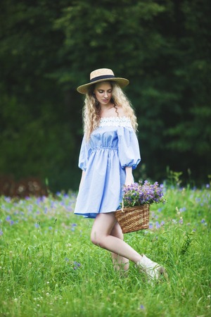Beautiful girl with basket of wild flowers. Woman with blond curly hair with a bouquet in a straw hat outdoors.の写真素材