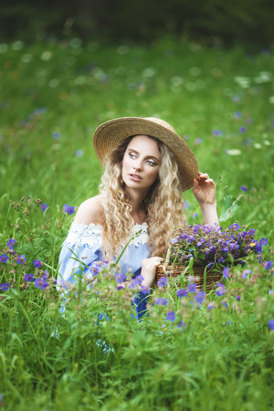 Beautiful girl with basket of wild flowers. Woman with blond curly hair with a bouquet in a straw hat outdoors.の写真素材
