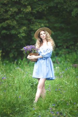 Beautiful girl with basket of wild flowers. Woman with blond curly hair with a bouquet in a straw hat outdoors.の写真素材