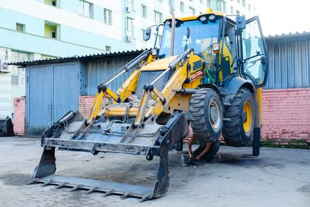 Backhoe loader stands leaning on a bucket. A man repairs a backhoe loader. Construction machinery in the city.の写真素材