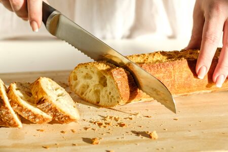 Slicing tasty, fresh, crisp bread. Beautiful female hands, a knife for cutting bread and sliced baguette closeup. Floury product on a cutting board. Healthy eating concept.の写真素材