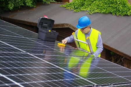 Installation and commissioning of solar panels. Renewable energy in a private home. Worker on ladder with electrical appliances on the roof. Ecological green power. Photovoltaic generator.の写真素材