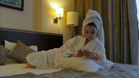 A young, beautiful woman in a white coat sits on a bed in a hotel, bedroom and writes an SMS message in the phone.の写真素材