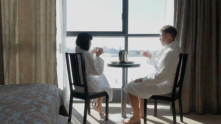 Young happy couple sitting by the window in hotel, at home. A woman and a man drink tea or coffee. A familyの写真素材